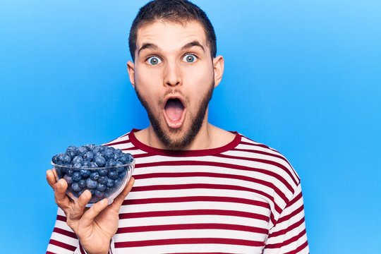 Young handsome man holding bowl with blueberries scared and amazed with open mouth for surprise, disbelief face