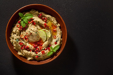 homemade spread of baked eggplant baba ganoush in a bowl with pomegranate seeds, lime, olive oil and lime slices on a black background