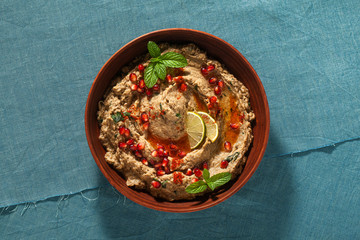 homemade spread of baked eggplant baba ganoush in a bowl with pomegranate seeds, lime, olive oil and lime slices on a blue linen tablecloth