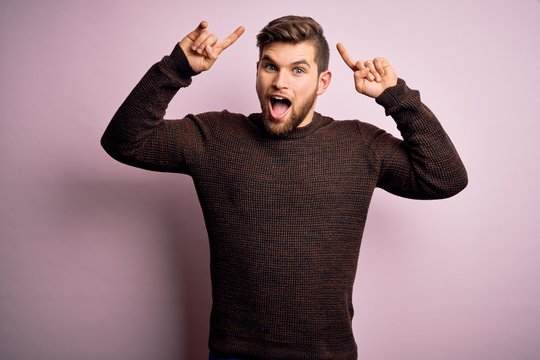 Young blond man with beard and blue eyes wearing casual sweater over pink background smiling amazed and surprised and pointing up with fingers and raised arms.