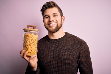Young blond man with beard and blue eyes holding bottle of Italian dry pasta macaroni with a happy...