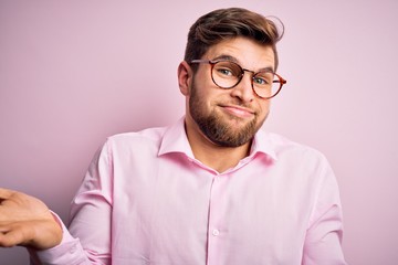 Young handsome blond man with beard and blue eyes wearing pink shirt and glasses clueless and...