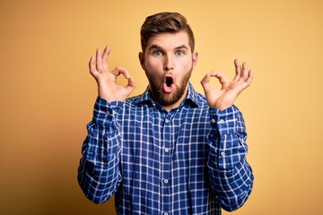 Young blond businessman with beard and blue eyes wearing shirt over yellow background looking surprised and shocked doing ok approval symbol with fingers. Crazy expression