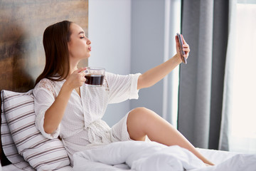 side view on woman having rest on bed, she looks at camera on smartphone, take photo. in domestic wear, at home