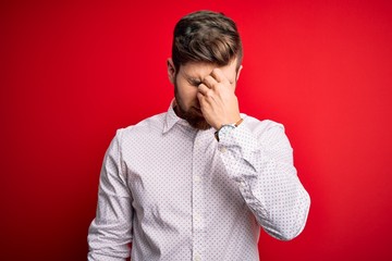 Young blond businessman with beard and blue eyes wearing elegant shirt over red background tired rubbing nose and eyes feeling fatigue and headache. Stress and frustration concept.