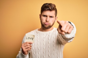 Young blond romantic man with beard and blue eyes holding paper with love message pointing with...