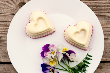 Souffle in the shape of a heart with cottage cheese, agar-agar and cream on a white plate on a wooden background.Violet flowers adorn the composition.Сoncept of healthy food and healthy dessert