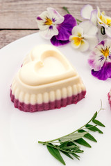 Souffle in the shape of a heart with cottage cheese, agar-agar and cream on a white plate on a wooden background.Edible flowers adorn the composition.Сoncept of healthy food and healthy dessert