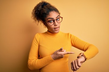 Young beautiful african american girl wearing sweater and glasses over yellow background In hurry pointing to watch time, impatience, upset and angry for deadline delay