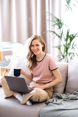 Smiling attractive woman in stylish casual wear works remotely at home sitting on sofa and drinking coffee. Distant work