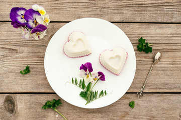 Souffle in the shape of a heart with cottage cheese, agar-agar and cream on a white plate on a wooden background.Violet flowers adorn the composition.Сoncept of healthy food and healthy dessert