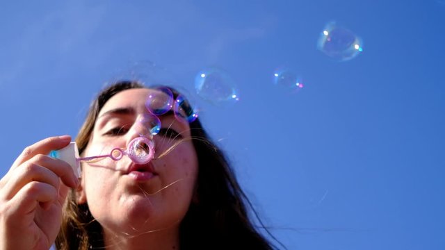 Young woman blowing soap bubbles towards the camera in slow motion. Bottom view of pretty auburn woman blowing soap bubbles on the roof