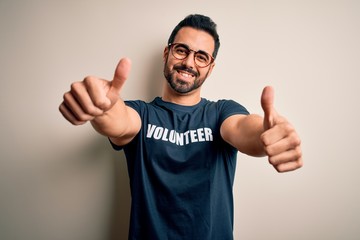 Handsome man with beard wearing t-shirt with volunteer message over white background approving doing positive gesture with hand, thumbs up smiling and happy for success. Winner gesture.