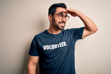 Handsome man with beard wearing t-shirt with volunteer message over white background very happy and smiling looking far away with hand over head. Searching concept.