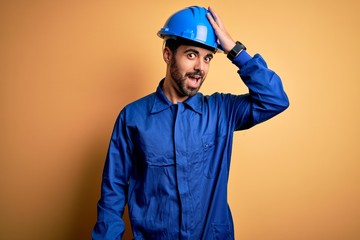 Mechanic man with beard wearing blue uniform and safety helmet over yellow background surprised with hand on head for mistake, remember error. Forgot, bad memory concept.
