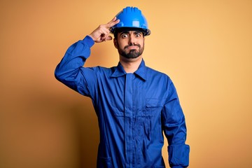 Mechanic man with beard wearing blue uniform and safety helmet over yellow background worried and stressed about a problem with hand on forehead, nervous and anxious for crisis