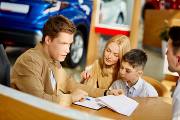 young caucasian family read documents and talk with consultant before purchase, they sit at table after view all cars represented there