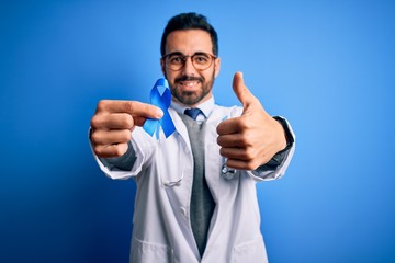 Young handsome doctor man with beard wearing stethoscope holding blue cancer ribbon happy with big smile doing ok sign, thumb up with fingers, excellent sign