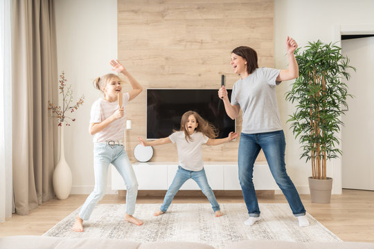 Happy Mother And Two Daughters Having Fun Singing Karaoke Song In Hairbrushes. Mother Laughing Enjoying Funny Lifestyle Activity With Teenage Girl At Home Together.