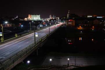 Fototapeta na wymiar Night city. The light trails on the street. Slow shutter speed photo. A long bridge across the river leads to the big city. A busy expressway in the town center.