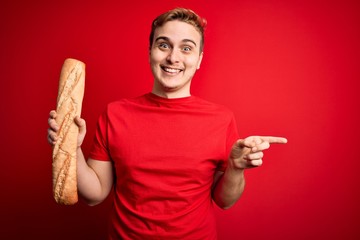 Young handsome redhead man holding fresh homemade bread over isolated red background Smiling happy pointing with hand and finger to the side