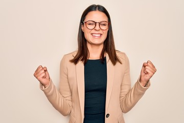 Young beautiful brunette businesswoman wearing jacket and glasses over white background excited for success with arms raised and eyes closed celebrating victory smiling. Winner concept.