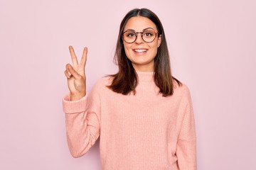 Young beautiful brunette woman wearing casual sweater and glasses over pink background showing and pointing up with fingers number two while smiling confident and happy.