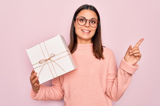 Young beautiful brunette woman holding birthday gift over isolated pink background Smiling happy pointing with hand and finger to the side