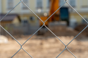 Excavator on a modern construction site. View of a construction site through a metal mesh fence. The fence in sharpness. Blurred background. Bokeh Soft focus.