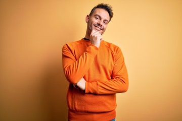 Young handsome man wearing orange casual sweater standing over isolated yellow background looking confident at the camera with smile with crossed arms and hand raised on chin. Thinking positive.