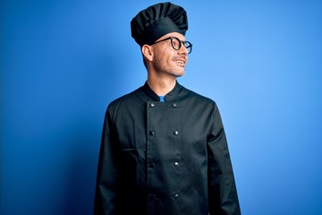 Young handsome chef man wearing cooker uniform and hat over isolated blue background looking away to side with smile on face, natural expression. Laughing confident.