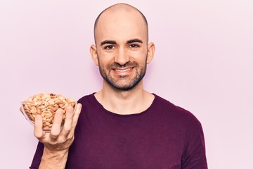 Young handsome bald man holding bowl with peanuts looking positive and happy standing and smiling with a confident smile showing teeth