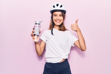 Young beautiful girl wearing bike helmet drinking bottle of water smiling happy and positive, thumb up doing excellent and approval sign