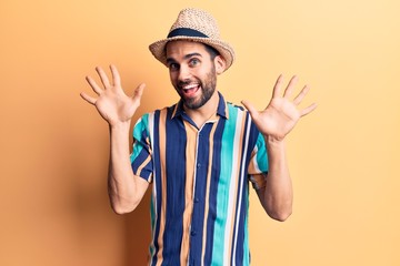 Young handsome man with beard wearing summer hat and shirt showing and pointing up with fingers number ten while smiling confident and happy.
