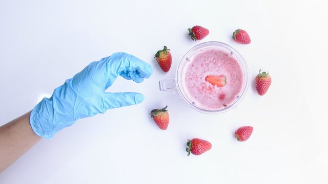 Vista superior de una mano con guante de protecci&oacute;n agarrando una jarra con batido de freza. Frezas sobre mesa blanca. Alimentaci&oacute;n saludable en la cuarentena por covid 19.  