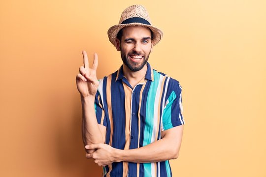 Young handsome man with beard wearing summer hat and shirt smiling with happy face winking at the camera doing victory sign. number two.