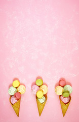 Summertime decorated border of ice cream cones filled with macarons and meringues on pink textured background, negative copy space. Stylish flat lay, top view minimalism creative layout. Vertical.