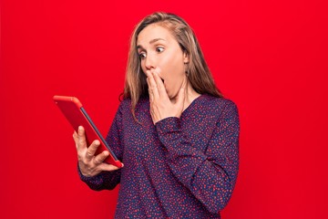 Young beautiful blonde woman using tablet over isolated red background covering mouth with hand, shocked and afraid for mistake. surprised expression