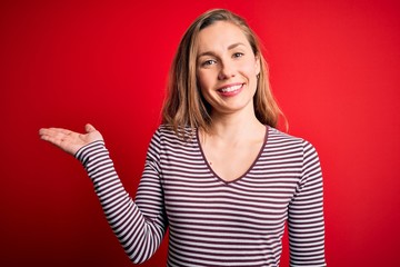 Young beautiful blonde woman wearing casual striped t-shirt over isolated red background smiling cheerful presenting and pointing with palm of hand looking at the camera.