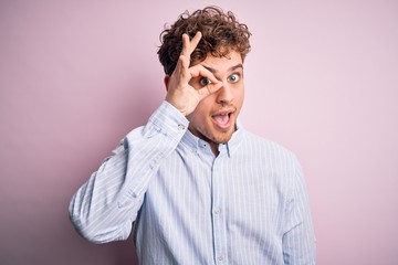 Young blond handsome man with curly hair wearing striped shirt over white background doing ok gesture with hand smiling, eye looking through fingers with happy face.