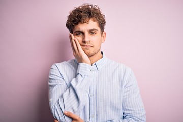 Young blond handsome man with curly hair wearing striped shirt over white background thinking looking tired and bored with depression problems with crossed arms.