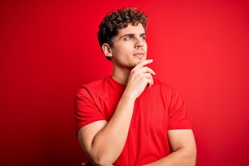 Young blond handsome man with curly hair wearing casual t-shirt over red background with hand on chin thinking about question, pensive expression. Smiling with thoughtful face. Doubt concept.