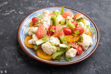 Vegetarian salad of cauliflower cabbage, kiwi, tomatoes, microgreen sprouts on black concrete background. Side view.