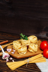 Kitchen cutting board, raw spaghetti, tagliatelle nests, tomatoes, garlic, ingredients for Italian dishes on a wooden background. Beautiful stylish workplace cook. Copy space for text