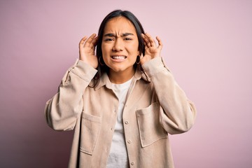 Young beautiful asian woman wearing casual shirt standing over pink background Trying to hear both hands on ear gesture, curious for gossip. Hearing problem, deaf