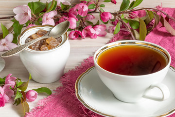 A Cup of tea, lump sugar in a white sugar bowl next to pink flowers on a white wooden table. Tea for Breakfast for the woman you love.