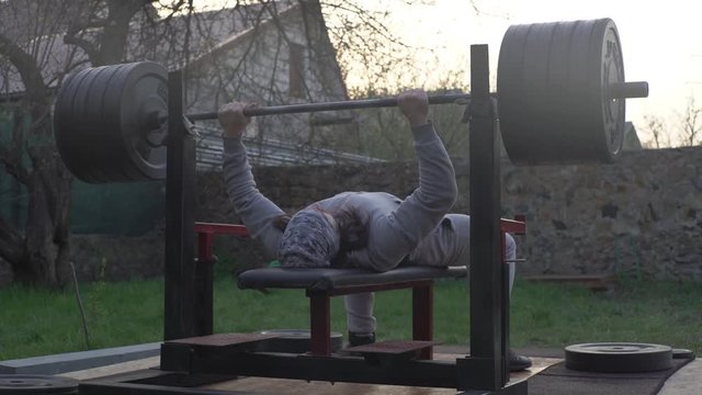 A man, an athlete, is training outdoors, doing chest press with a barbell