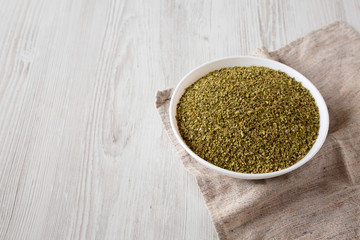 Dried Green Greek Oregano Spice in a white bowl on a white wooden background, high angle view. Copy space.
