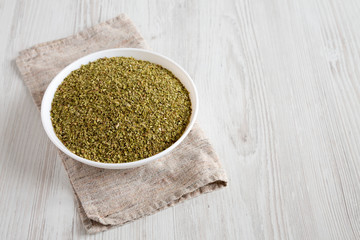Dried Green Greek Oregano Spice in a white bowl on a white wooden surface, high angle view. Copy space.