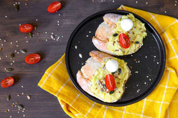 Two sandwiches with avocado and cherry tomatoes and mozzarella with seeds of flax, sesame, pumpkin and half-sunflower in a black ceramic plate on a wooden table with a yellow napkin. Flat lay.